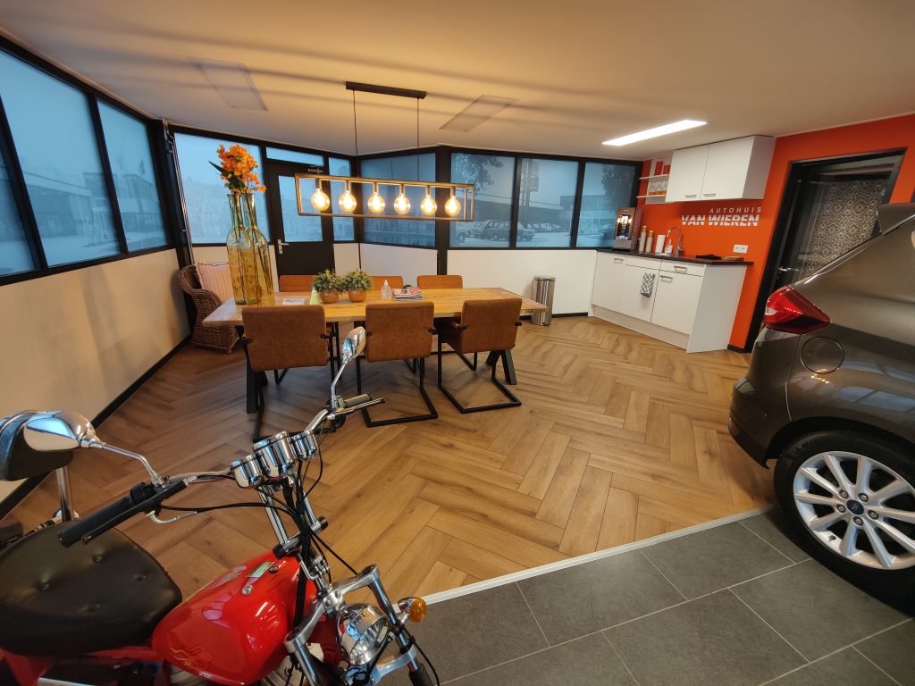 De koffiehoek in de showroom met een houten tafel, hanglampen en de oranje wand met het logo